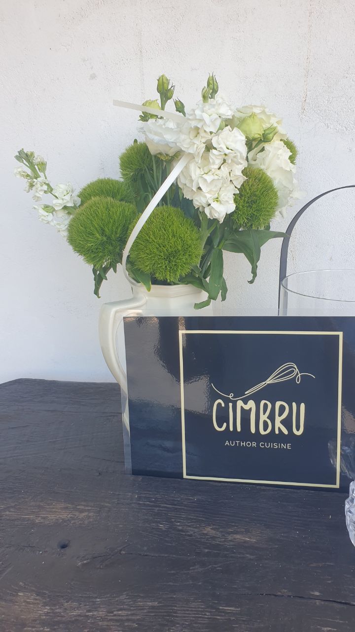 A bouquet of white flowers and green pom-pom blooms beside a Cimbru Author Cuisine sign on a dark table